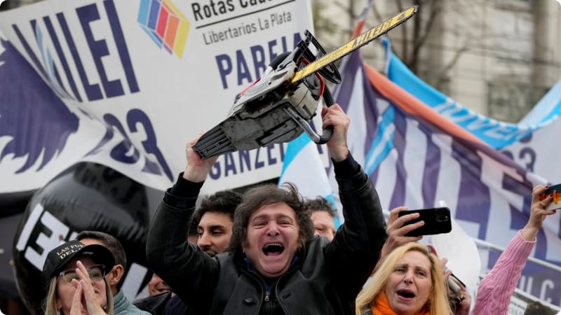 A man joyfully holds a chainsaw aloft in a crowd during a protest, with various banners and flags in the background.
