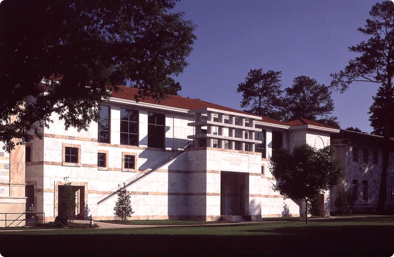 A modernist building with a white and beige stone facade, featuring large windows and a distinctive staircase, surrounded by trees and grass.