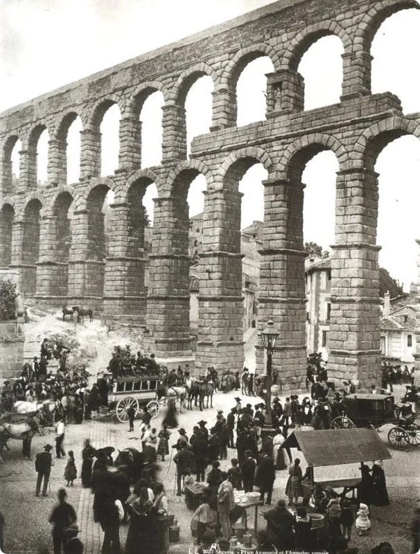 A historic black-and-white photograph of a large Roman aqueduct with multiple arches, surrounded by a bustling crowd, horse-drawn carriages, and market stalls.