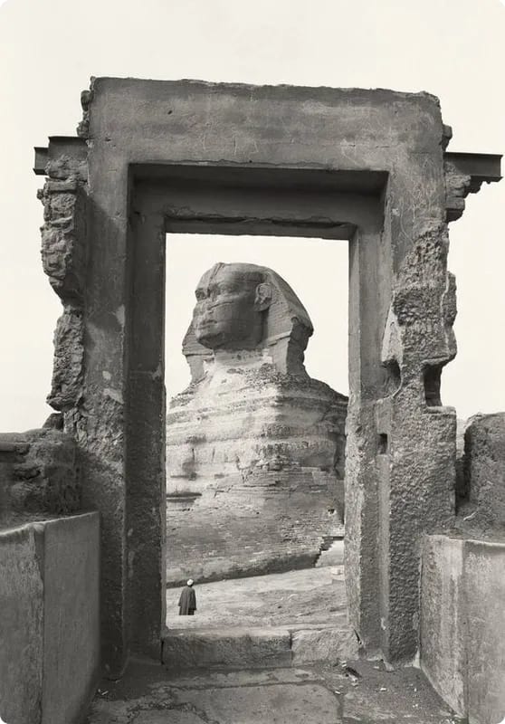 A black and white photograph of the Great Sphinx of Giza viewed through a stone archway, with a person standing in the foreground.