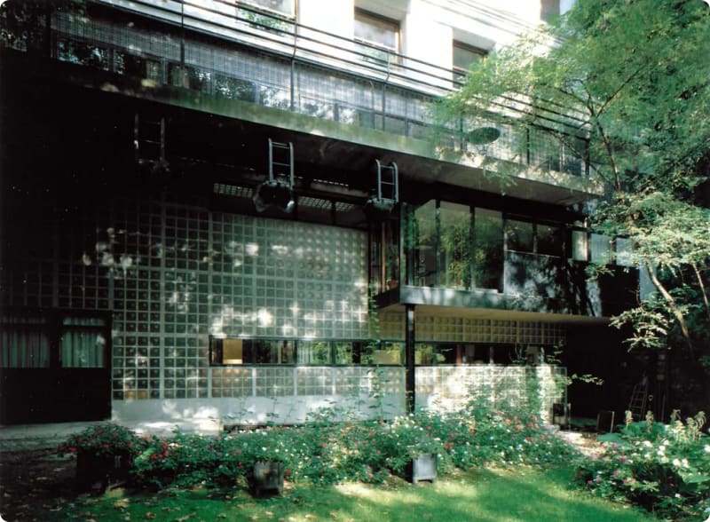 A modern building featuring a glass block facade, large windows, and a balcony, surrounded by greenery and flowers.