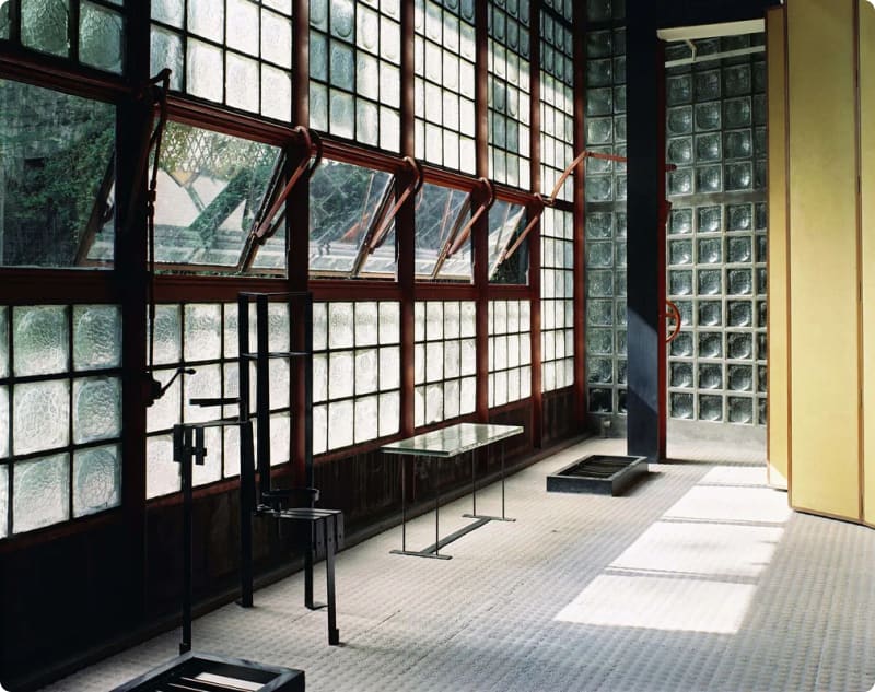 Interior of a modern room featuring large glass block windows, a glass table, and minimalist furniture, with natural light streaming in.