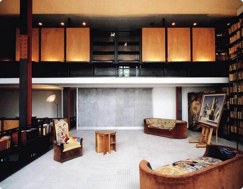 A spacious modern interior featuring a mix of wooden and black shelving, two unique sofas, a small wooden table, and a large wall with a gray surface.