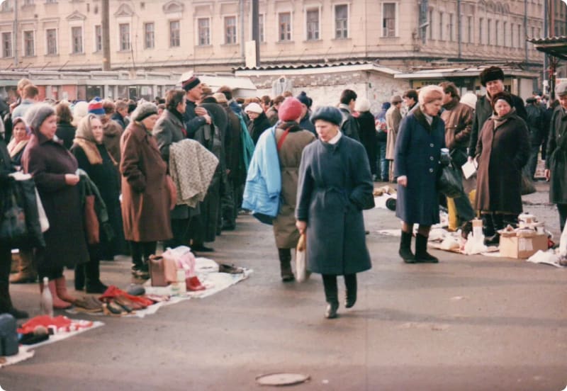 A crowded outdoor market scene with people in winter clothing, some standing in line and others browsing items laid out on the ground.
