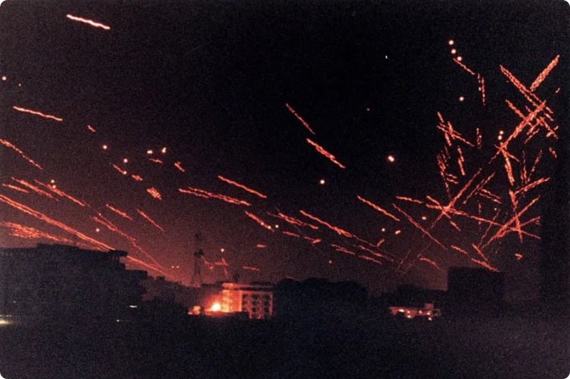 A night sky filled with streaks of red light, resembling flares or fireworks, above silhouetted buildings.