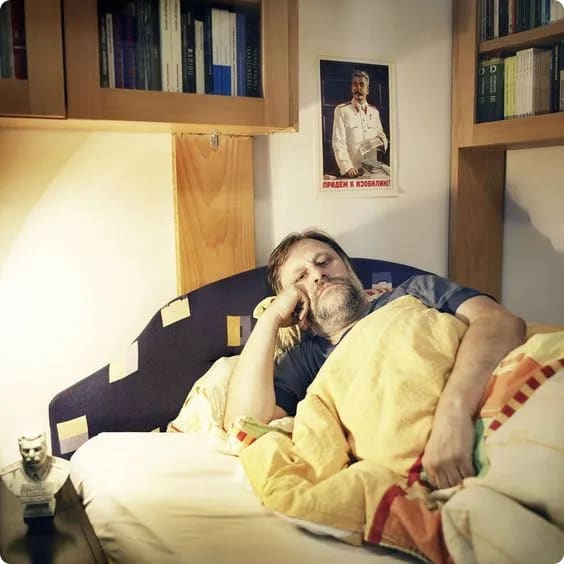 A man with a beard is sleeping in bed, covered with a colorful blanket, with a poster of a historical figure on the wall behind him and bookshelves above.