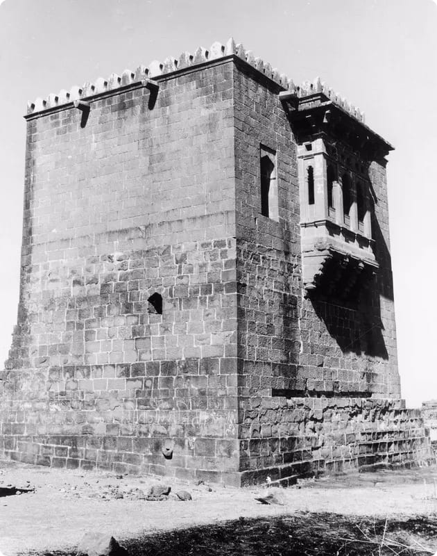 A black and white photograph of a stone tower with a flat roof and decorative elements, featuring a small balcony and windows, set against a clear sky.