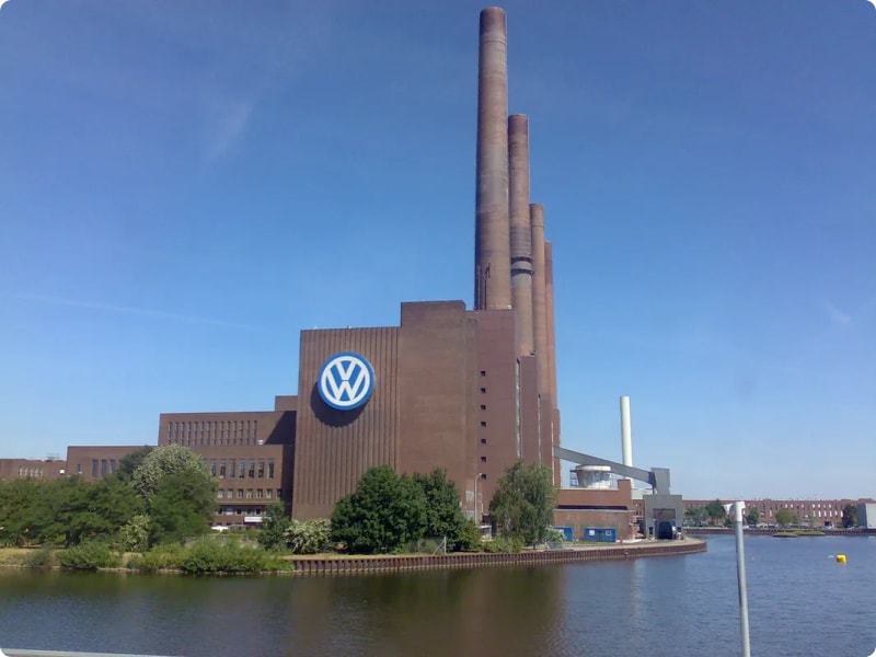 Volkswagen factory with tall smokestacks beside a body of water under a clear blue sky.