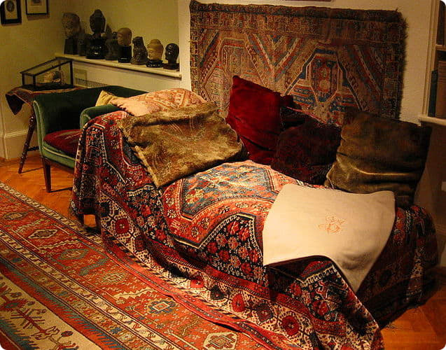 A cozy seating area featuring a couch draped with colorful patterned textiles and cushions, surrounded by a decorative rug and a shelf with sculptures.