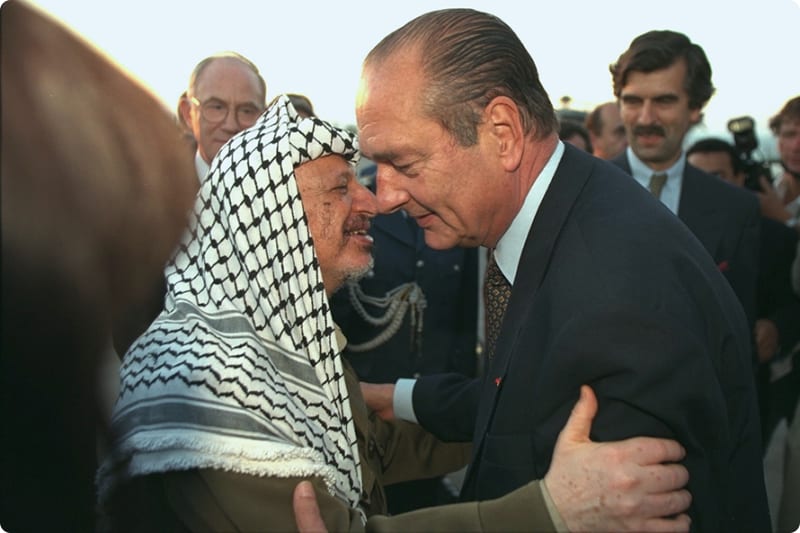 A close embrace between two men, one wearing a traditional Palestinian keffiyeh and the other in a suit, with a group of people in the background.