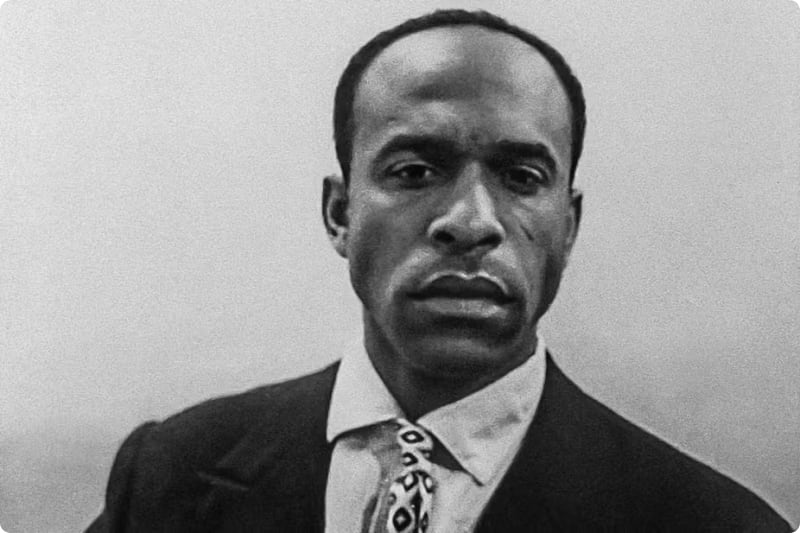 Black and white portrait of a man in a suit with a patterned tie, looking directly at the camera.