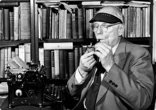 An elderly man wearing glasses and a visor is sitting at a typewriter, smoking a pipe, with a bookshelf filled with books in the background.