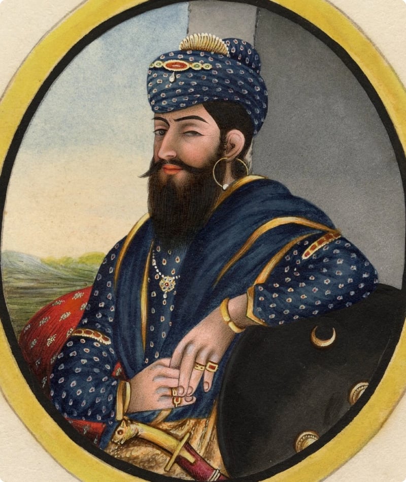 A regal man in traditional attire, adorned with jewelry and a turban, sits confidently with a sword at his side, set against a scenic background.