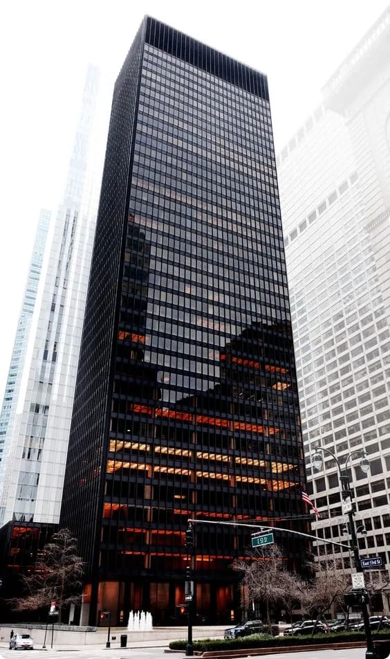 A tall, black glass skyscraper surrounded by other buildings, with visible reflections and illuminated interior lights.