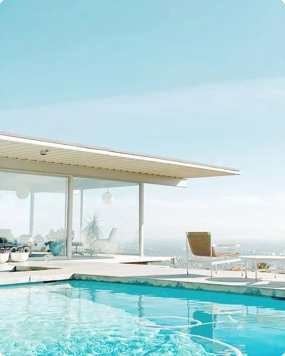 A modern glass house with a flat roof overlooking a swimming pool and a scenic view under a clear blue sky.