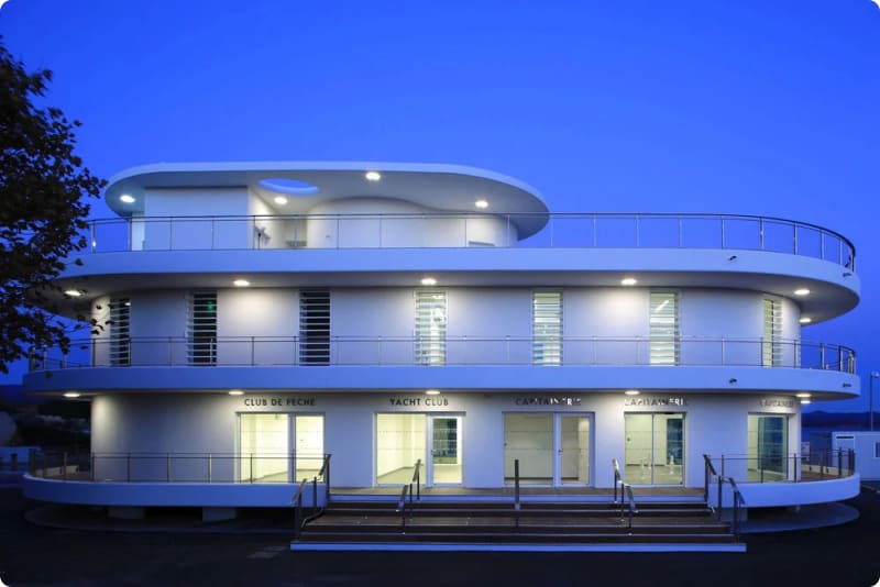 Modern white building with curved architecture, featuring multiple balconies and illuminated signage for a yacht club, set against a twilight sky.