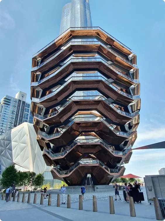 The Vessel, a honeycomb-like structure made of bronze and glass, stands prominently in Hudson Yards, New York City, with people gathered around its base.