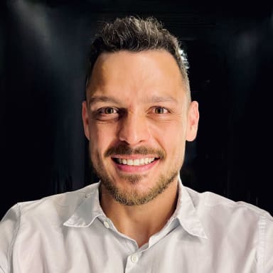 A headshot of Weverson Mamédio smiling. He has short, dark hair styled upwards, a light stubble beard, and clear skin. He is wearing a white button-up shirt with an open collar. The background is dark, which highlights his face as the focal point. His warm and confident smile suggests approachability and professionalism.