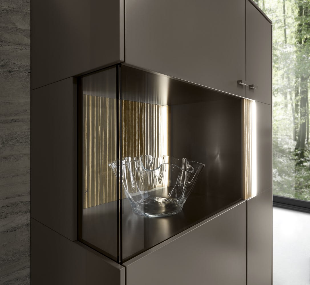 La combinaison de vie SALE 25 Linee, un bol en verre, présenté dans un meuble moderne avec portes vitrées et intérieur éclairé, sur fond de vue sur la forêt. La combinaison de vie SALE 25 Linee, un bol en verre, présenté dans un meuble moderne avec portes vitrées et intérieur éclairé, sur fond de vue sur la forêt.