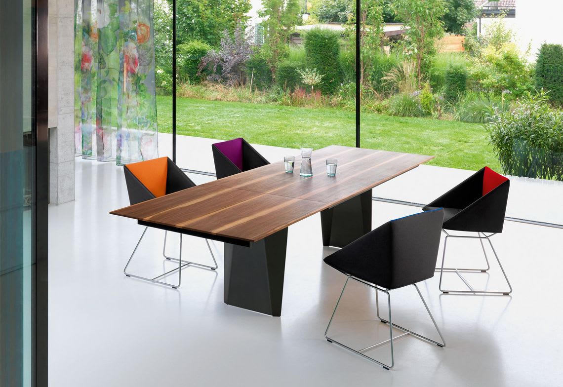 Le Girsberger Tisch Bela est en combinaison avec des chaises colorées dans une pièce avec des murs en verre et offre une vue sur un jardin vert luxuriant. Le Girsberger Tisch Bela est en combinaison avec des chaises colorées dans une pièce avec des murs en verre et offre une vue sur un jardin vert luxuriant.