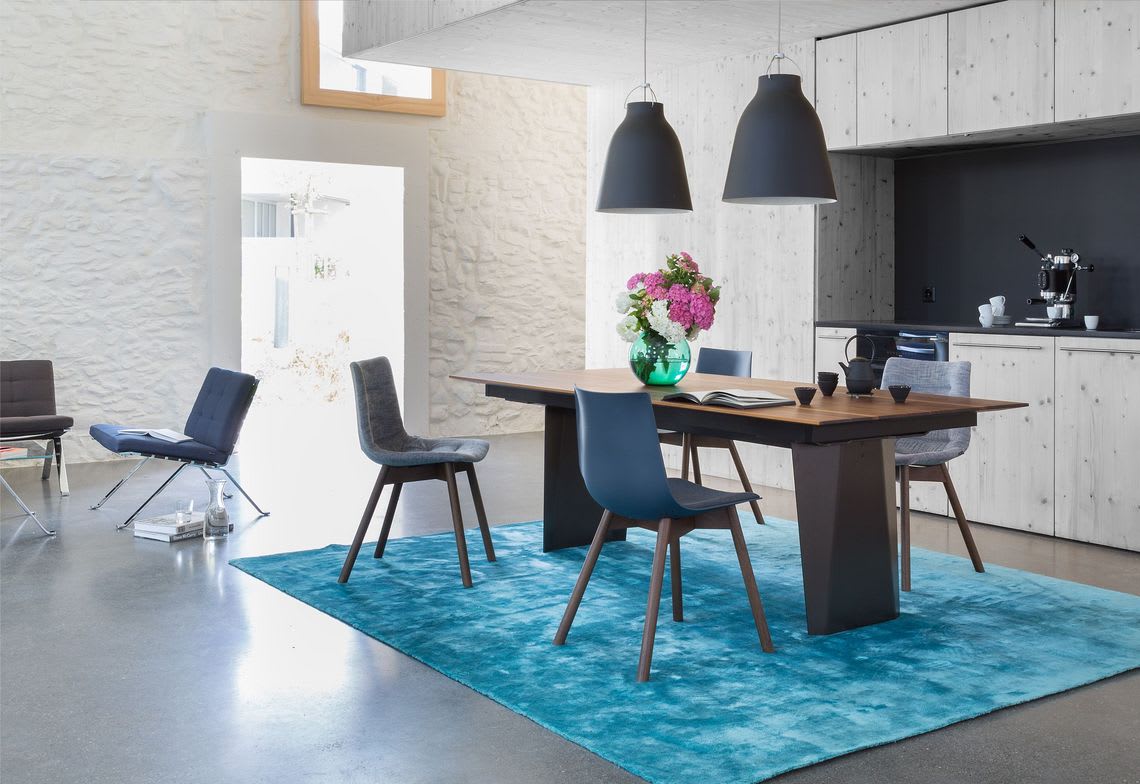 Une salle à manger moderne avec la table en bois Girsberger Tisch Bela, des chaises bleues, des suspensions et un tapis turquoise. Design léger et minimaliste. Une salle à manger moderne avec la table en bois Girsberger Tisch Bela, des chaises bleues, des suspensions et un tapis turquoise. Design léger et minimaliste.