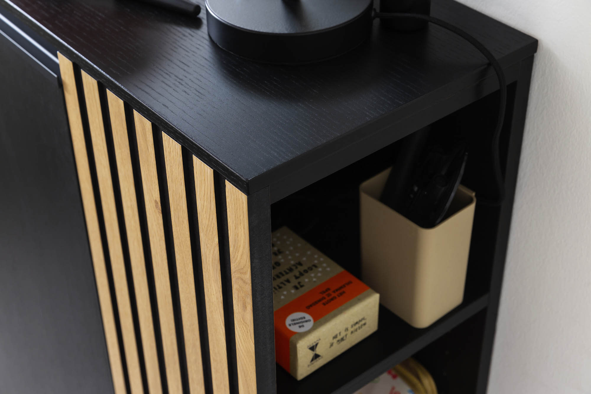 Die Garderobe Punta besteht aus einem schwarzen Rahmen mit hellen Holzlatten, in dem Bücher und ein beiger Behälter mit einer Fernbedienung auf der unteren Ablage stehen. Die Garderobe Punta besteht aus einem schwarzen Rahmen mit hellen Holzlatten, in dem Bücher und ein beiger Behälter mit einer Fernbedienung auf der unteren Ablage stehen.