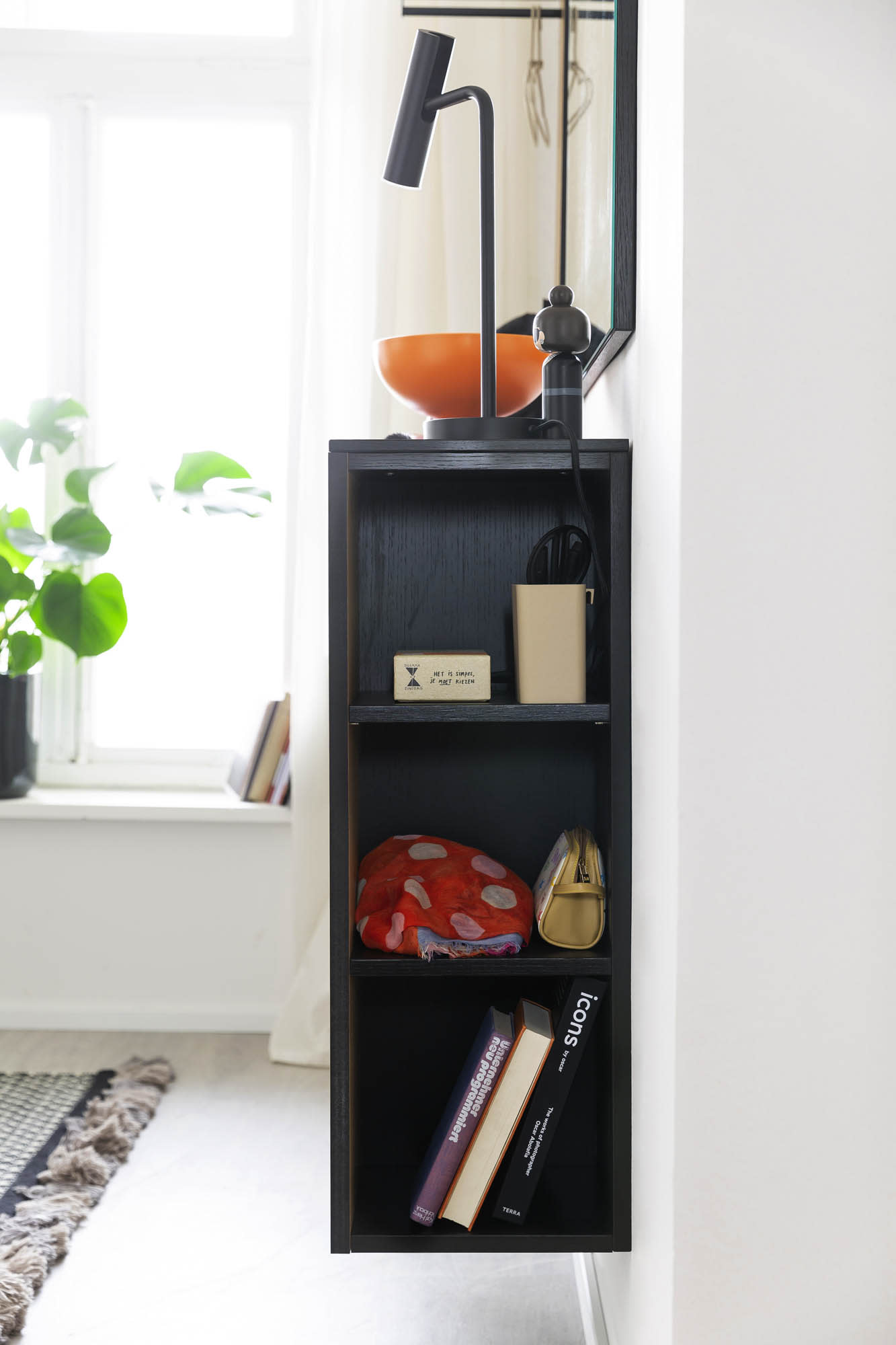 Die Garderobe Punta, ein kompaktes schwarzes Regal, bietet Platz für Bücher, eine Lampe und Dekoration an einer weißen Wand in der Nähe eines Fensters. Die Garderobe Punta, ein kompaktes schwarzes Regal, bietet Platz für Bücher, eine Lampe und Dekoration an einer weißen Wand in der Nähe eines Fensters.