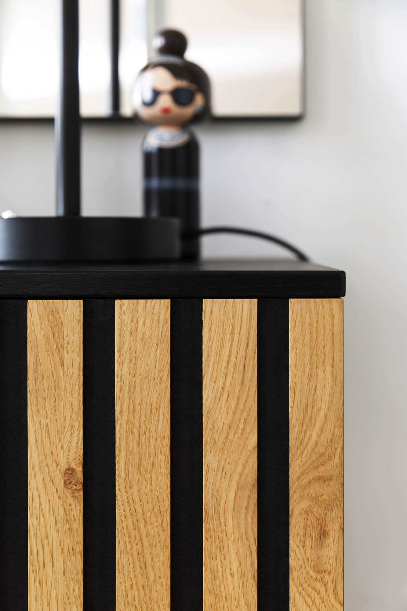 Nahaufnahme des Holzschranks Garderobe Punta mit vertikalen schwarzen Streifen; im Hintergrund erscheint eine verschwommene Puppenfigur mit Sonnenbrille. Nahaufnahme des Holzschranks Garderobe Punta mit vertikalen schwarzen Streifen; im Hintergrund erscheint eine verschwommene Puppenfigur mit Sonnenbrille.