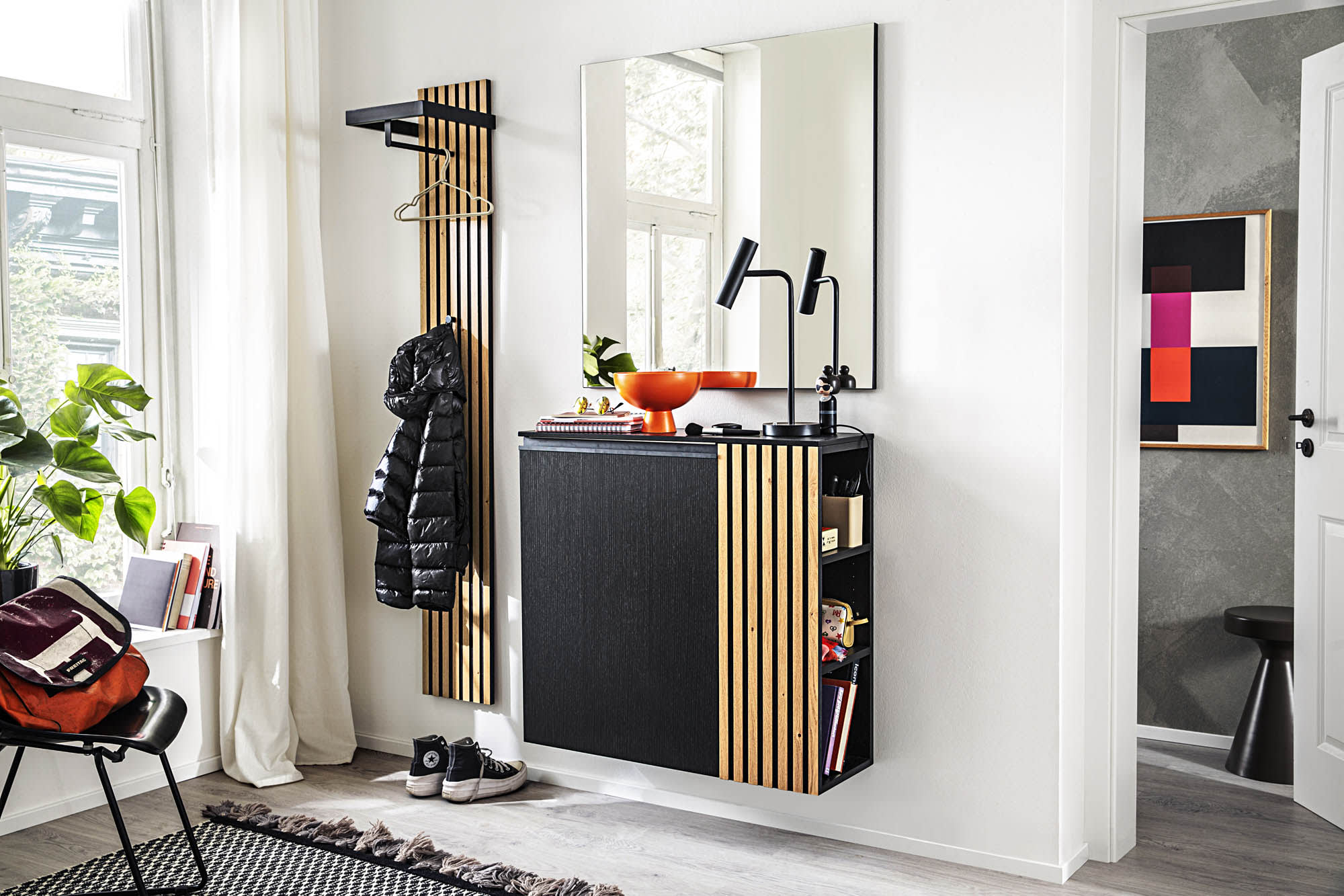 Moderner Eingangsbereich mit der Garderobe Punta in Schwarz und Holz, ergänzt durch einen Spiegel, einen Garderobenständer, Pflanzen und bunte dekorative Akzente. Moderner Eingangsbereich mit der Garderobe Punta in Schwarz und Holz, ergänzt durch einen Spiegel, einen Garderobenständer, Pflanzen und bunte dekorative Akzente.
