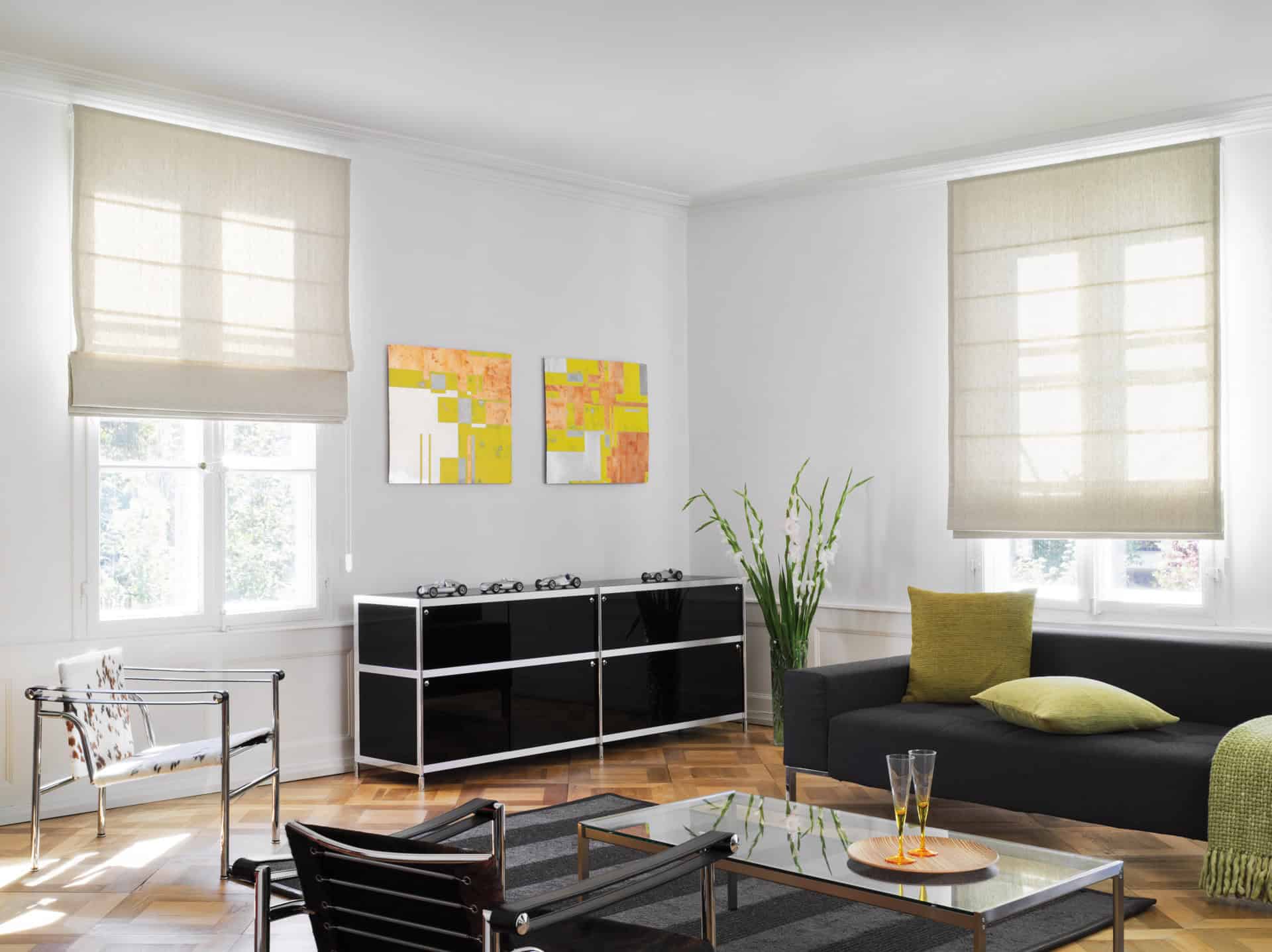 Salon moderne avec meubles noirs, art abstrait et grandes fenêtres avec stores romains beiges.