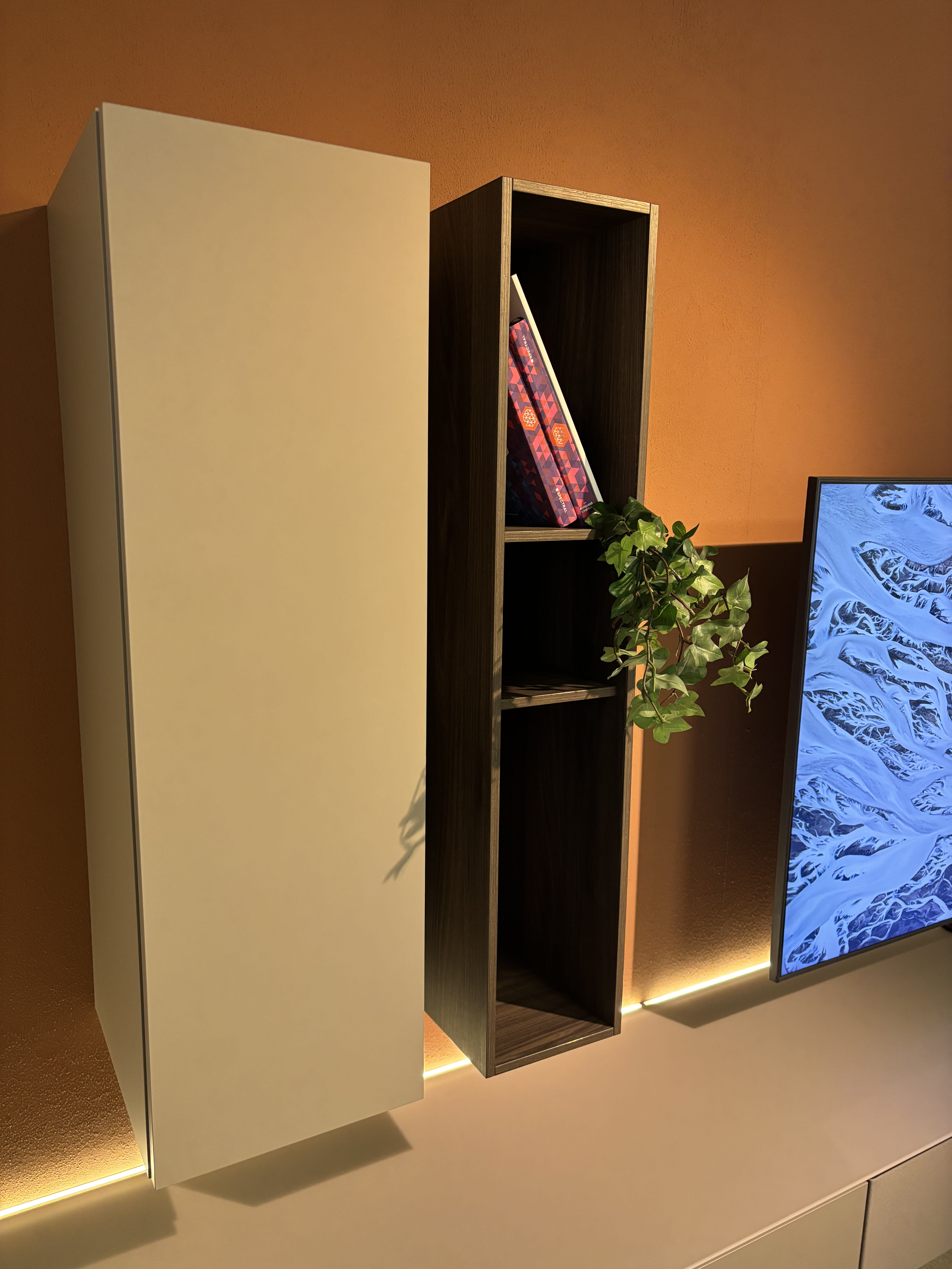 Das TV-Möbel Next von Spectral ist mit modernen Wandregalen mit Büchern und einer Topfpflanze ausgestattet, die neben einem Fernseher an einer orangefarbenen Wand stehen, die von einer Akzentbeleuchtung beleuchtet wird.