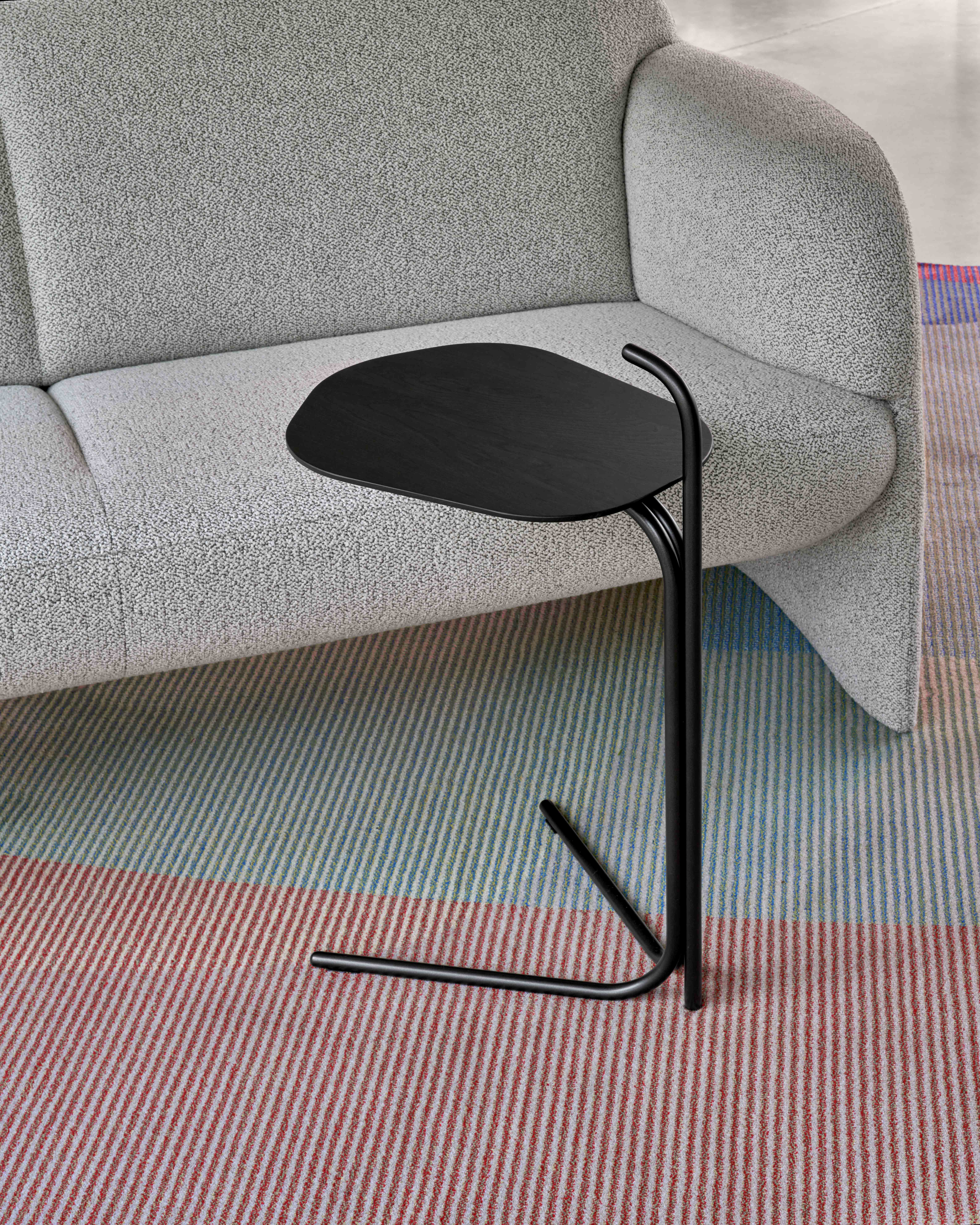 Ein kleiner schwarzer Beistelltisch steht neben dem Leolux-Sofa Modius, das mit grauem Strukturstoff bezogen ist, auf einem bunt gestreiften Teppich. Ein kleiner schwarzer Beistelltisch steht neben dem Leolux-Sofa Modius, das mit grauem Strukturstoff bezogen ist, auf einem bunt gestreiften Teppich.