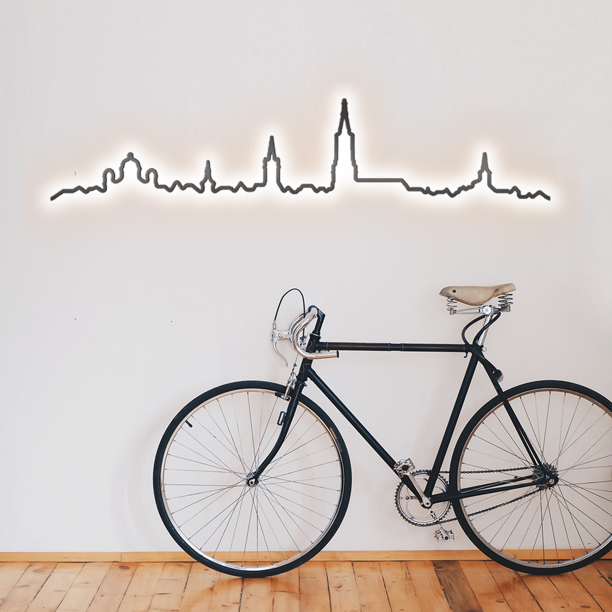 Ein schwarzes Velo lehnt an einer weissen Wand, gekrönt vom beleuchteten Kontura City Bern Skyline Dekor. Ein schwarzes Velo lehnt an einer weissen Wand, gekrönt vom beleuchteten Kontura City Bern Skyline Dekor.