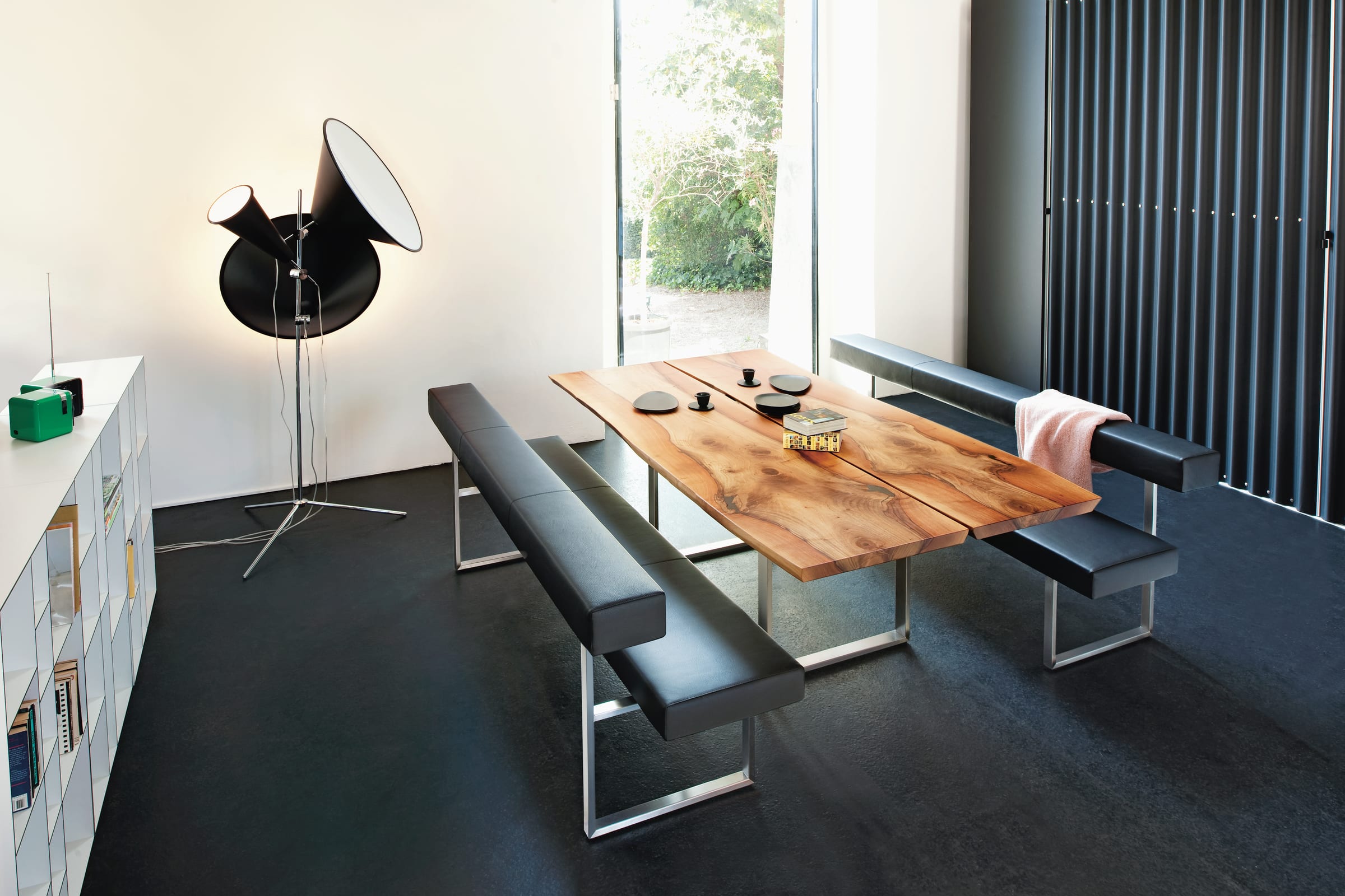 Moderner Essbereich mit Holztisch, schwarzen Sitzbänken der Girsberger Sitzbank Permesso, Stehlampe und einem Fenster, das Tageslicht einlässt. Moderner Essbereich mit Holztisch, schwarzen Sitzbänken der Girsberger Sitzbank Permesso, Stehlampe und einem Fenster, das Tageslicht einlässt.