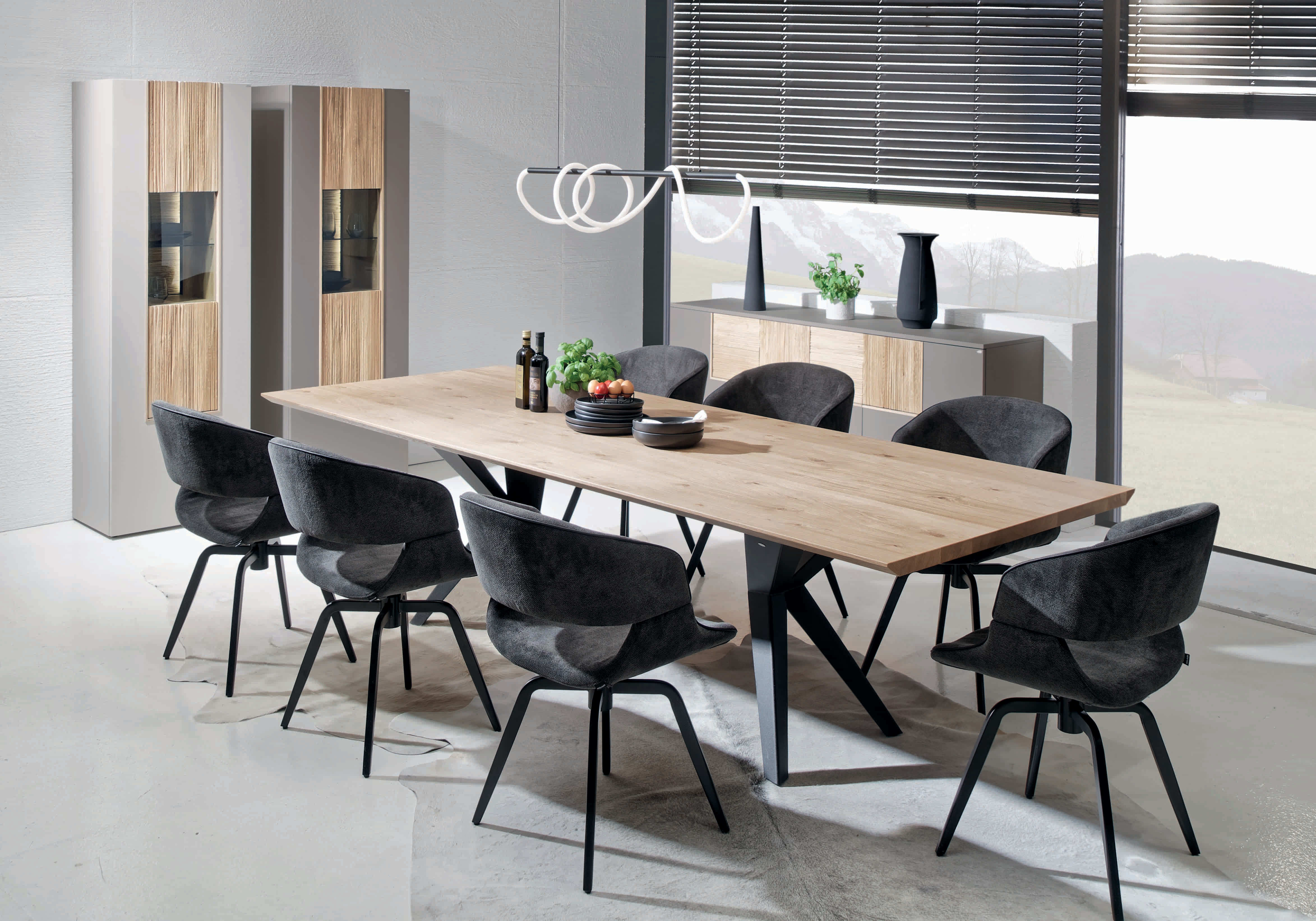 Modernes Esszimmer mit dem Voglauer Lacera Tisch, acht schwarzen Stühlen und minimalistischem Dekor in der Nähe großer Fenster.