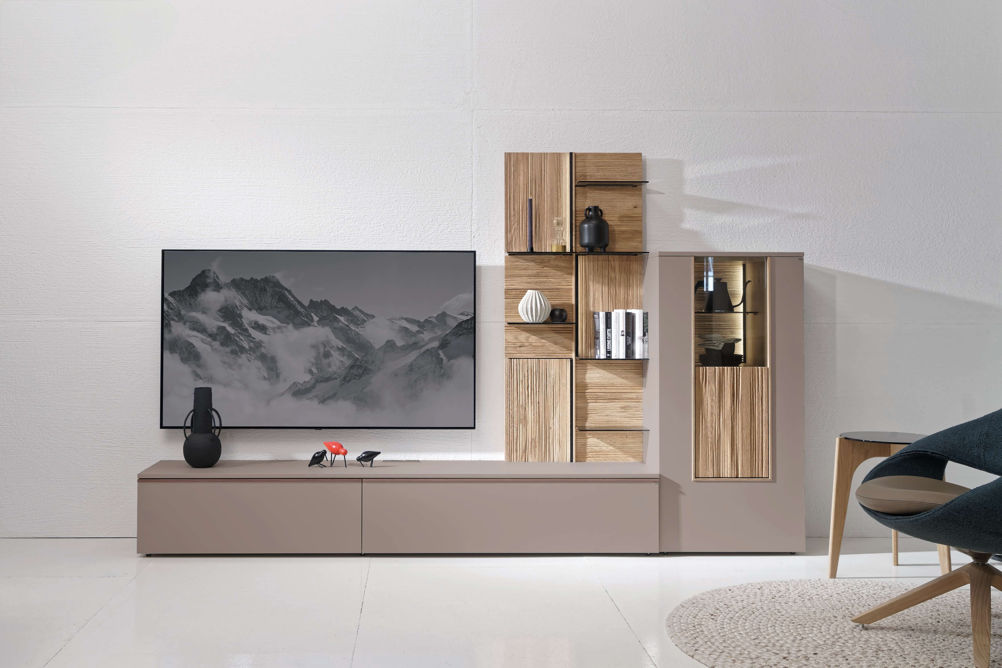 Modernes Wohnzimmer mit dem Voglauer Wohnmöbel Lacera: ein schlichter Schrank mit einem an der Wand befestigten Fernseher, stilvollen Regalen, Büchern und dekorativen Vasen.