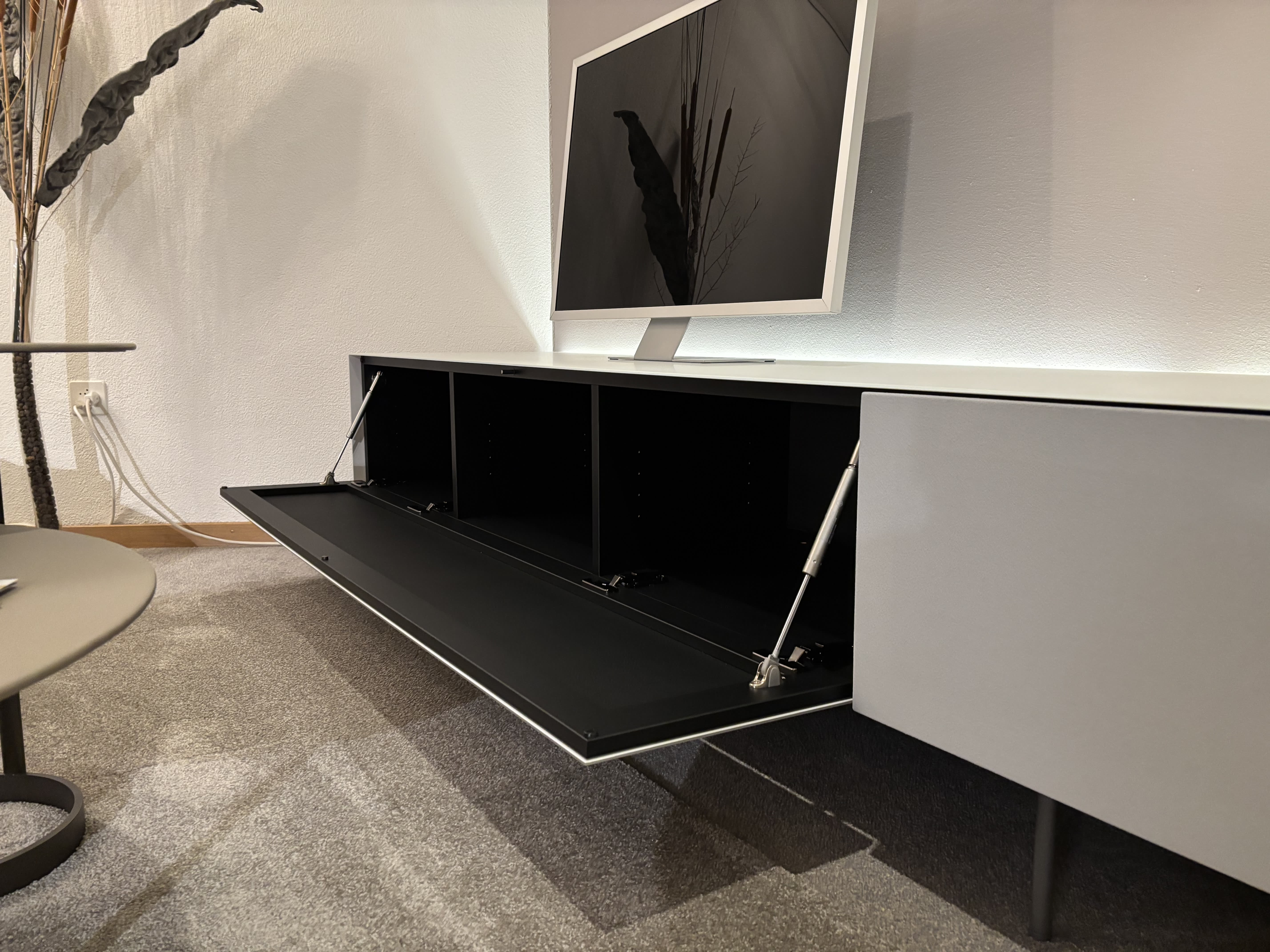 Modernes Wohnzimmer mit einem Flachbildfernseher auf einem Büromöbel mit 3 Boxenfenstern, einem eleganten weißen Schrank mit offenem Stauraum, alles auf einem grauen Teppich. Modernes Wohnzimmer mit einem Flachbildfernseher auf einem Büromöbel mit 3 Boxenfenstern, einem eleganten weißen Schrank mit offenem Stauraum, alles auf einem grauen Teppich.