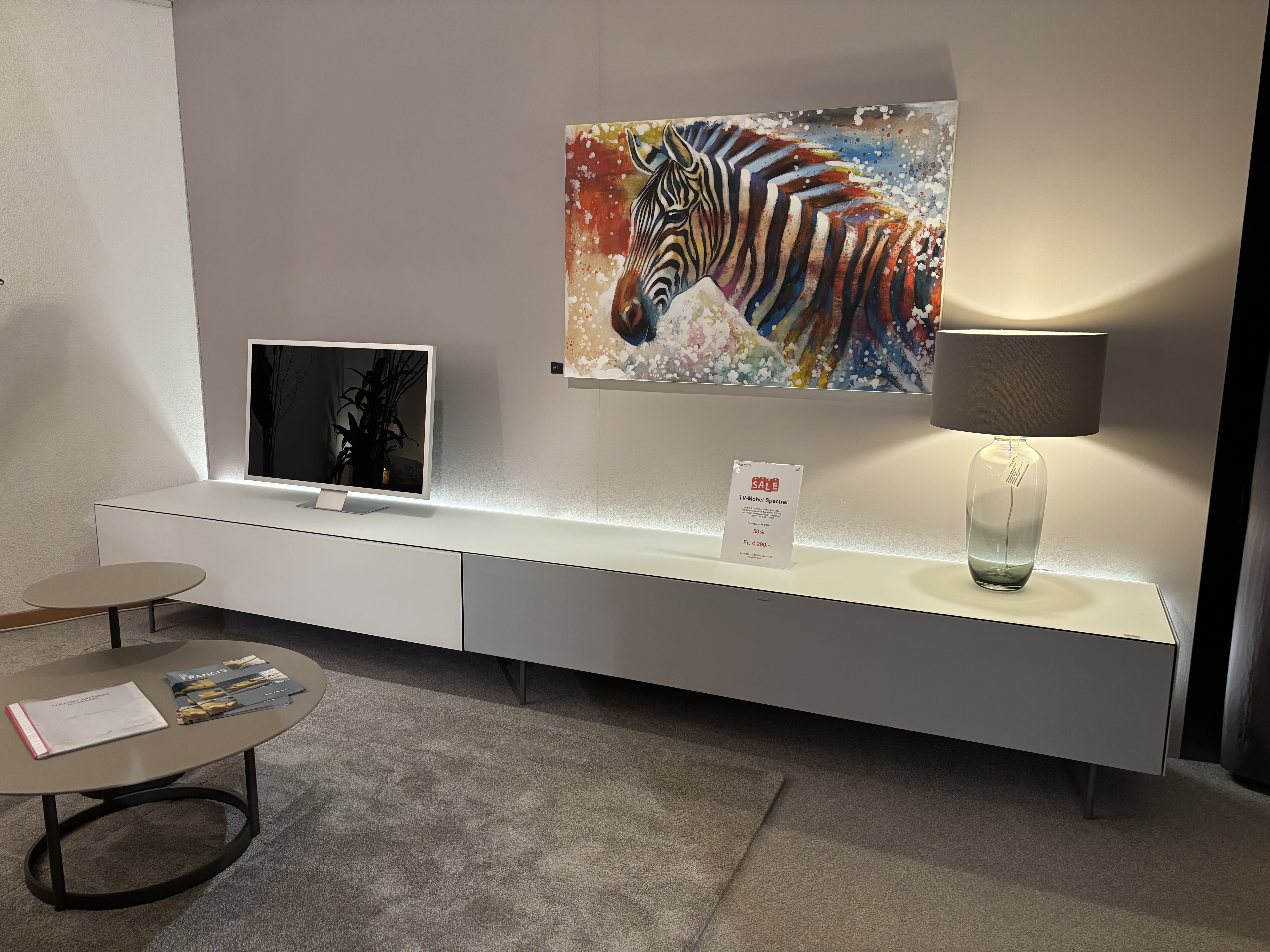 Moderne Wohnzimmereinrichtung mit Fernseher, Zebragemälde, Lampe und runden Couchtischen auf einem grauen Teppich mit Büromöbeln mit 3 Boxenfenstern. Moderne Wohnzimmereinrichtung mit Fernseher, Zebragemälde, Lampe und runden Couchtischen auf einem grauen Teppich mit Büromöbeln mit 3 Boxenfenstern.