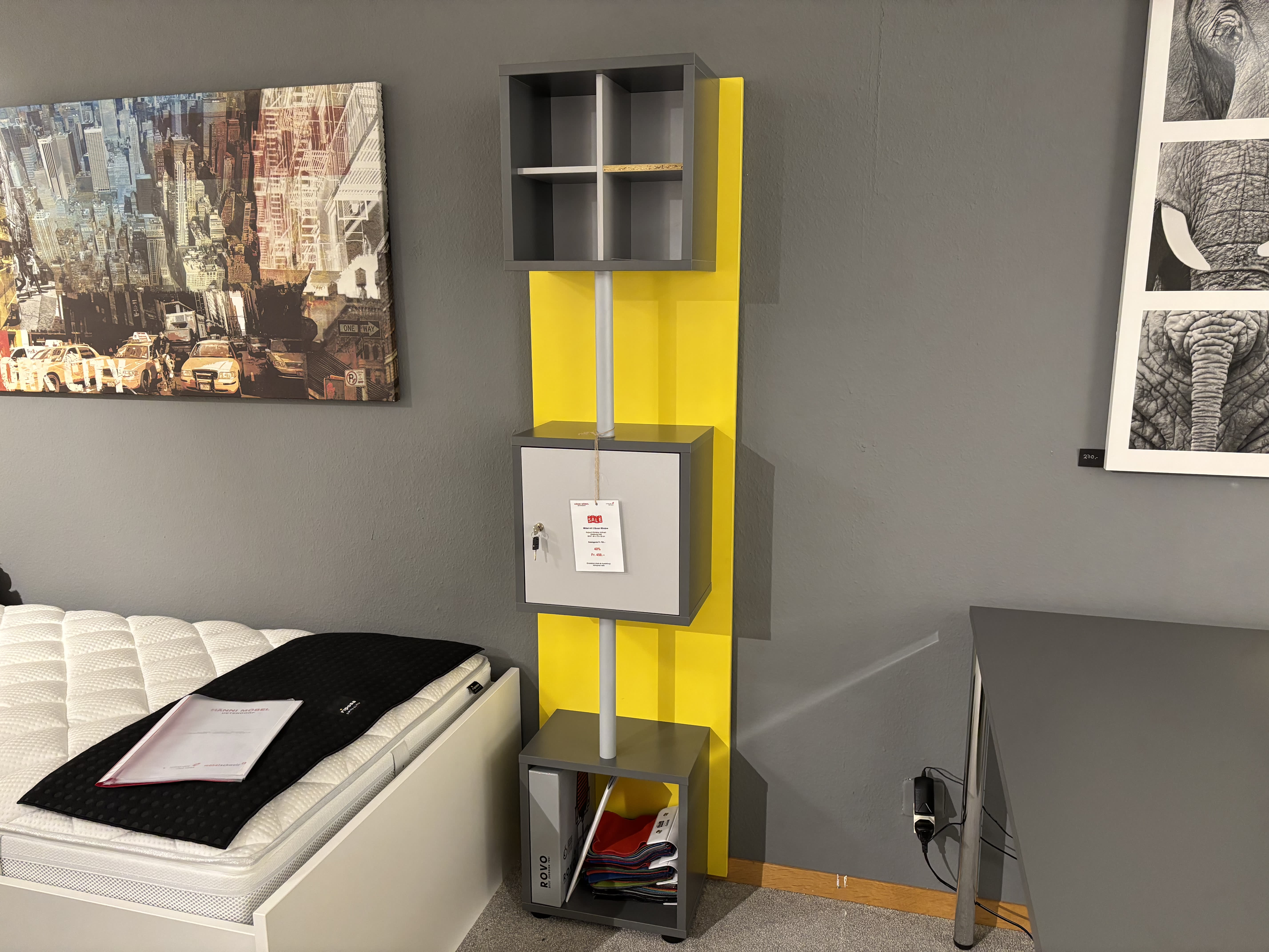 Das Büromöbel mit 3 Boxen Window, ein hohes Regal in Grau und Gelb, steht neben einem Bett mit weißer Matratze und schwarzer Decke. Das Büromöbel mit 3 Boxen Window, ein hohes Regal in Grau und Gelb, steht neben einem Bett mit weißer Matratze und schwarzer Decke.