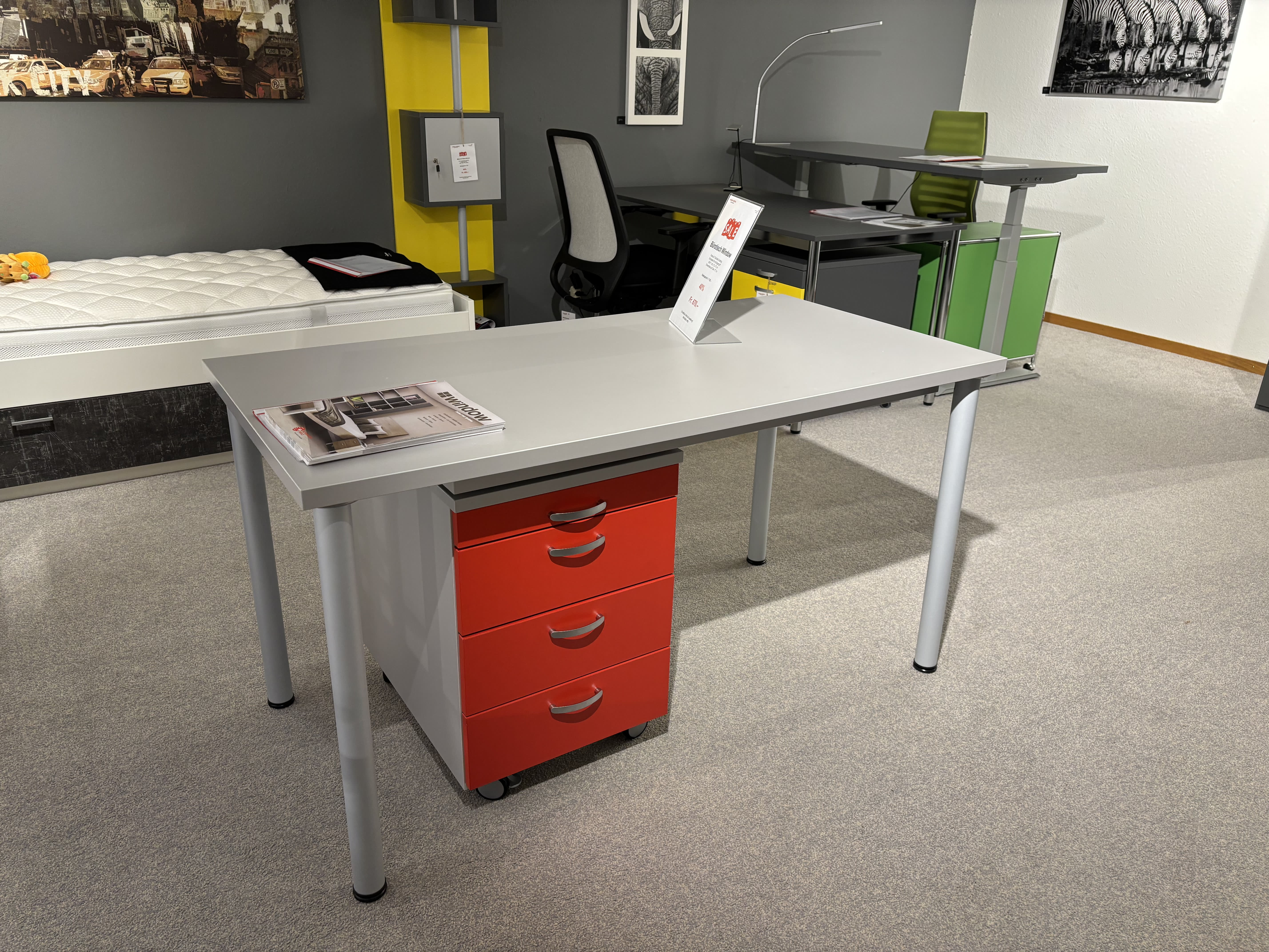 Zeitgenössischer Showroom mit Büromöbeln mit 3 Boxen Window mit grauem Schreibtisch, roten Schubladen, Stuhl und bunten Regalen. Teppichboden und eine Matratze in der Nähe runden das Ambiente ab. Zeitgenössischer Showroom mit Büromöbeln mit 3 Boxen Window mit grauem Schreibtisch, roten Schubladen, Stuhl und bunten Regalen. Teppichboden und eine Matratze in der Nähe runden das Ambiente ab.