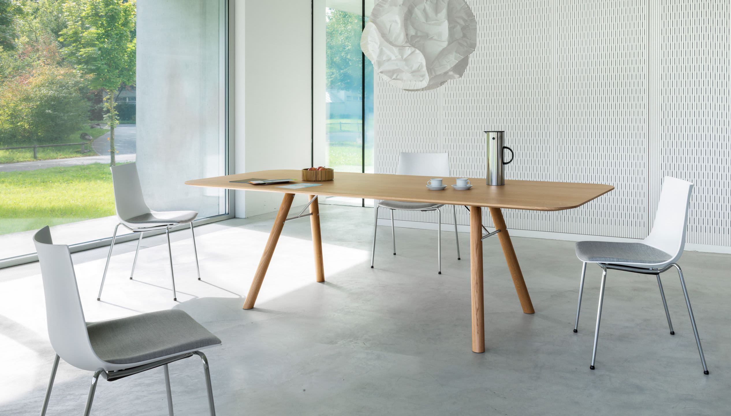 Minimalistischer Konferenzraum mit dem Holztisch Esstisch Akio, vier weißen Stühlen und großen Fenstern mit Blick ins Grüne. Minimalistischer Konferenzraum mit dem Holztisch Esstisch Akio, vier weißen Stühlen und großen Fenstern mit Blick ins Grüne.