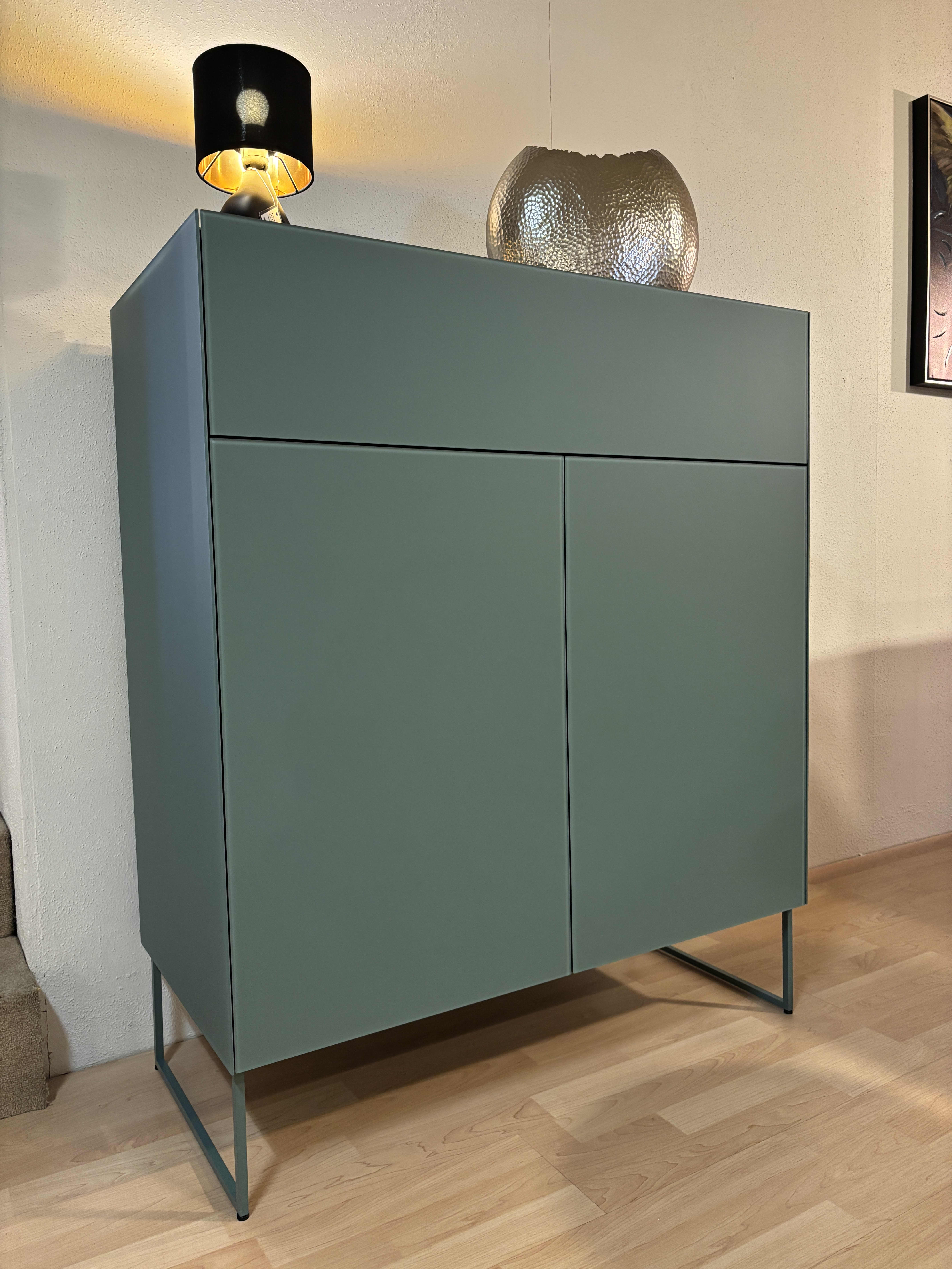 Das Spectral Sideboard 101cm zeichnet sich durch ein minimalistisches grünes Design mit Metallbeinen aus, das durch eine schwarze Lampe und eine Schale aus strukturiertem Glas ergänzt wird, die auf einem Holzboden stehen. Das Spectral Sideboard 101cm zeichnet sich durch ein minimalistisches grünes Design mit Metallbeinen aus, das durch eine schwarze Lampe und eine Schale aus strukturiertem Glas ergänzt wird, die auf einem Holzboden stehen.