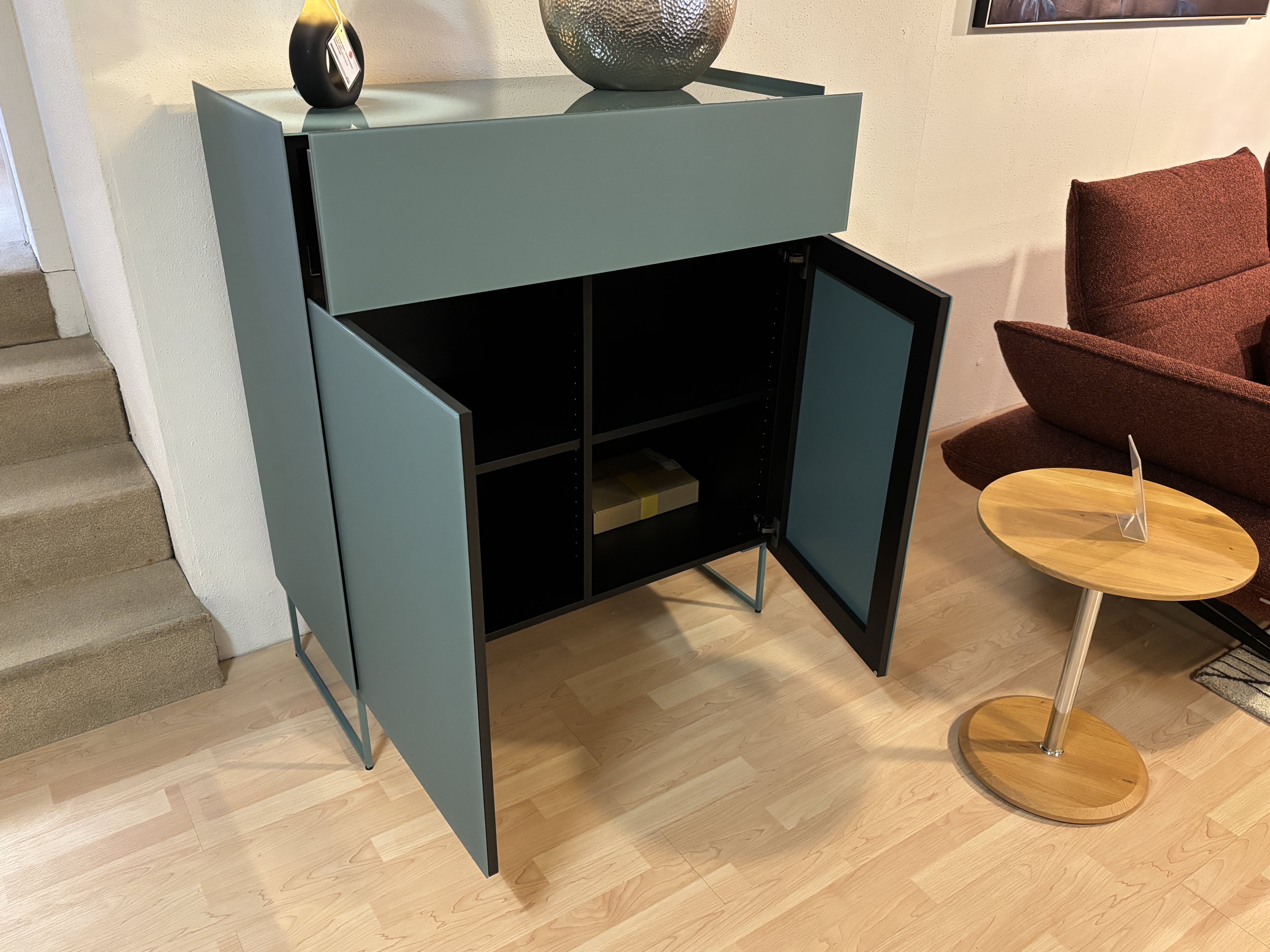 Das Spectral Sideboard 101cm in modernem Teal steht mit geöffneten Türen neben einem runden Holztisch und gibt den Blick frei auf Innenböden und eine Box. Das Spectral Sideboard 101cm in modernem Teal steht mit geöffneten Türen neben einem runden Holztisch und gibt den Blick frei auf Innenböden und eine Box.