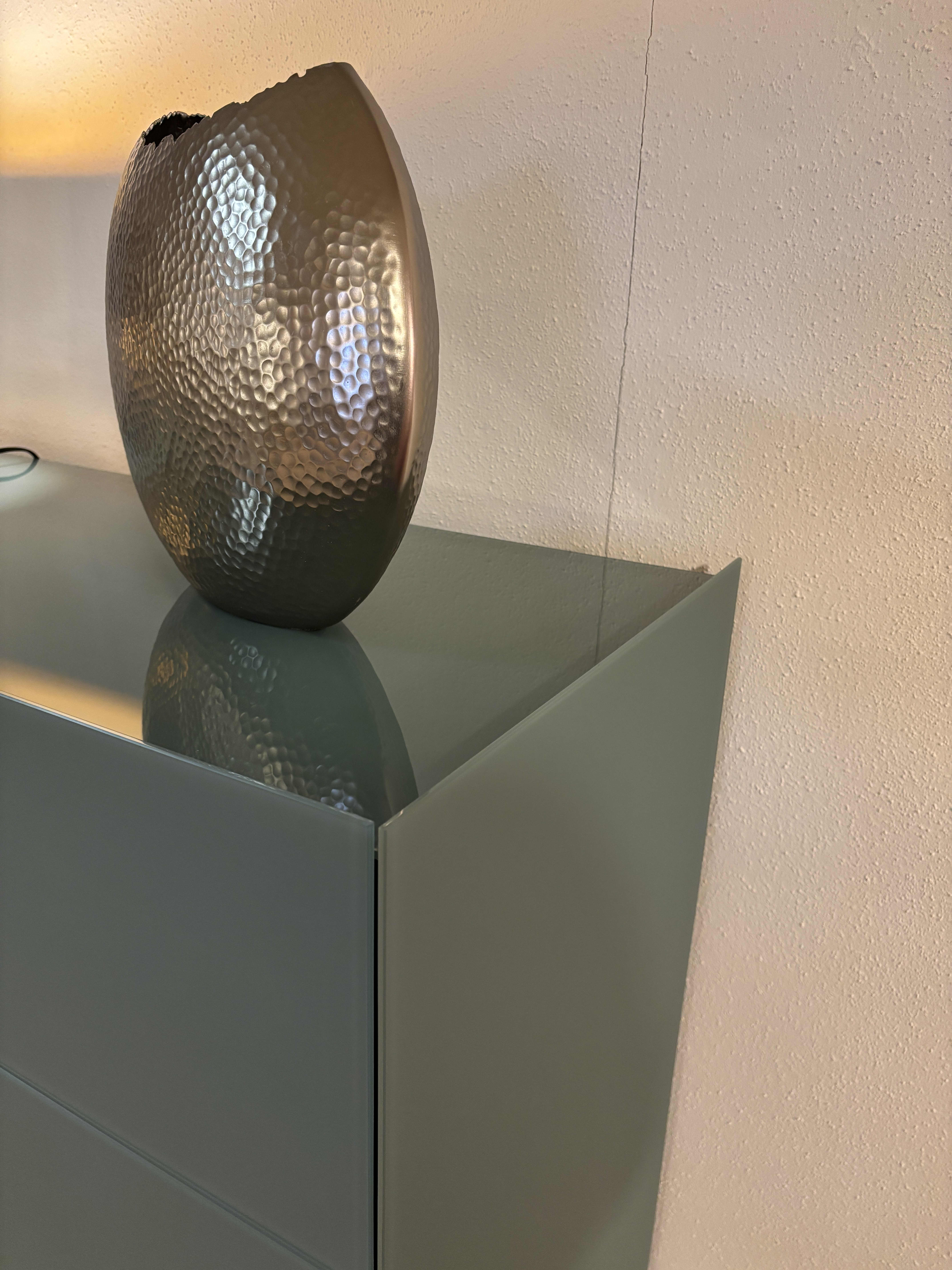 Eine metallische, strukturierte Vase steht auf dem Spectral Sideboard 101cm, einem glänzenden, hellgrünen Schrank, vor einer beigen Wand. Eine metallische, strukturierte Vase steht auf dem Spectral Sideboard 101cm, einem glänzenden, hellgrünen Schrank, vor einer beigen Wand.