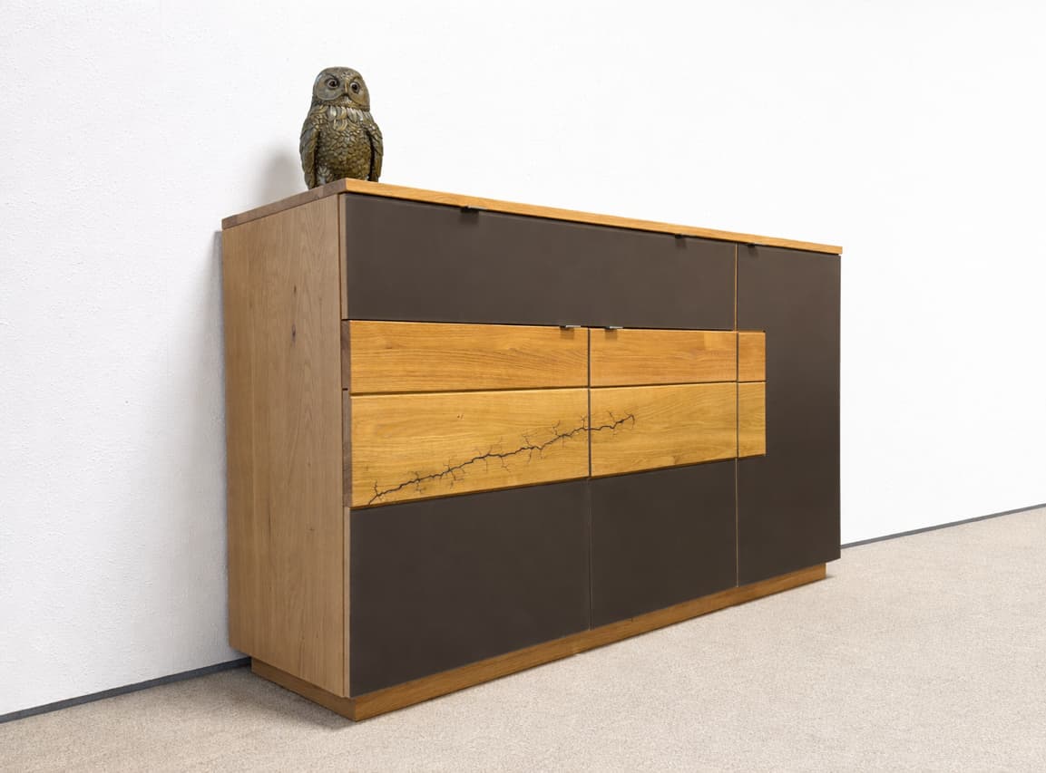 Le buffet Wimmer Signatura se distingue par son design moderne en bois avec des panneaux sombres et est couronné d'une sculpture de chouette placée devant un mur blanc. Le buffet Wimmer Signatura se distingue par son design moderne en bois avec des panneaux sombres et est couronné d'une sculpture de chouette placée devant un mur blanc.