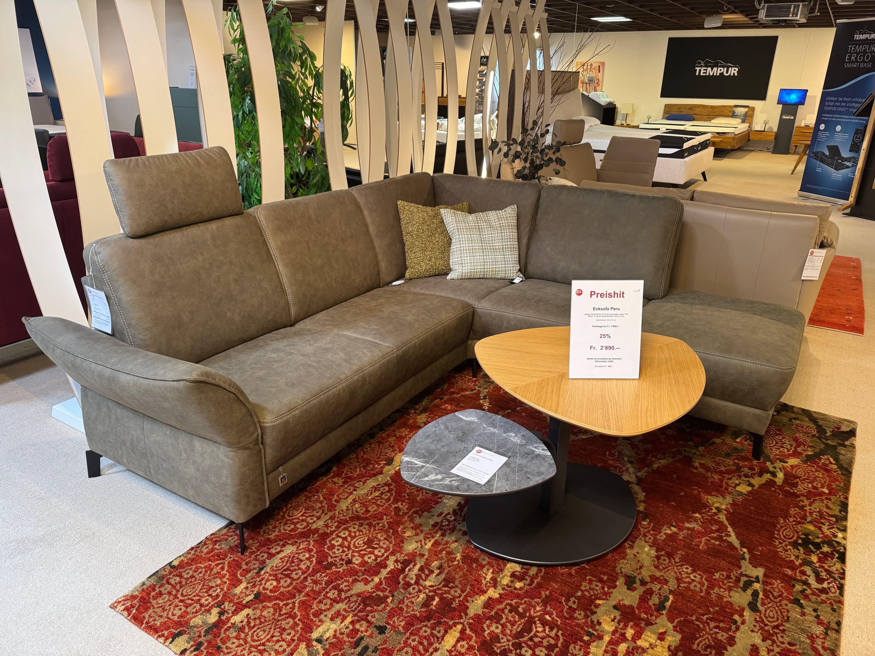 Das Ponsel Sofa Peru, ein graues Sektionssofa mit Wurfkissen, steht neben einem hölzernen Couchtisch auf einem rot gemusterten Teppich im Ausstellungsraum. Das Ponsel Sofa Peru, ein graues Sektionssofa mit Wurfkissen, steht neben einem hölzernen Couchtisch auf einem rot gemusterten Teppich im Ausstellungsraum.