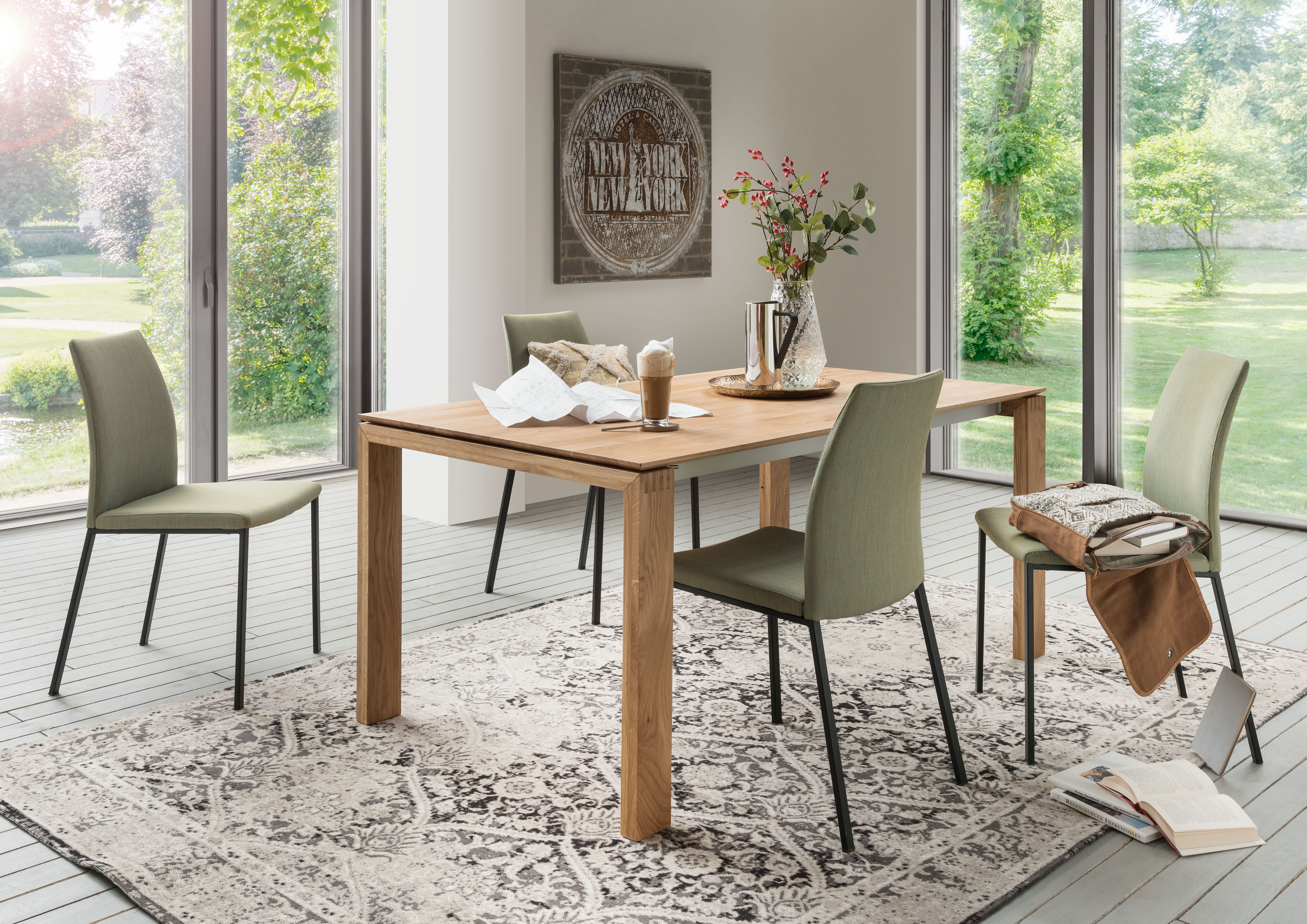 Modernes Esszimmer mit dem Holztisch Sit Mobilia Tisch Merkur Allegro, vier grünen Stühlen und großen Fenstern mit Blick auf einen üppigen Garten.