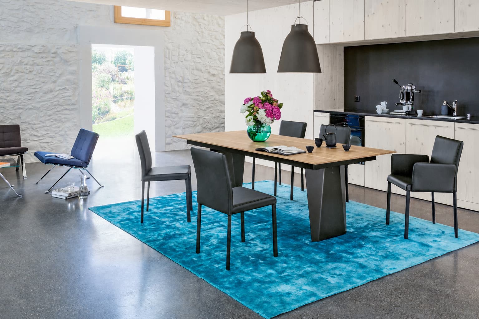 Moderne Essecke mit sechs schwarzen Stühlen von Girsberger Stuhl Kuba, einem Holztisch, Pendelleuchten, einem blauen Teppich und einer Blumenvase. Moderne Essecke mit sechs schwarzen Stühlen von Girsberger Stuhl Kuba, einem Holztisch, Pendelleuchten, einem blauen Teppich und einer Blumenvase.