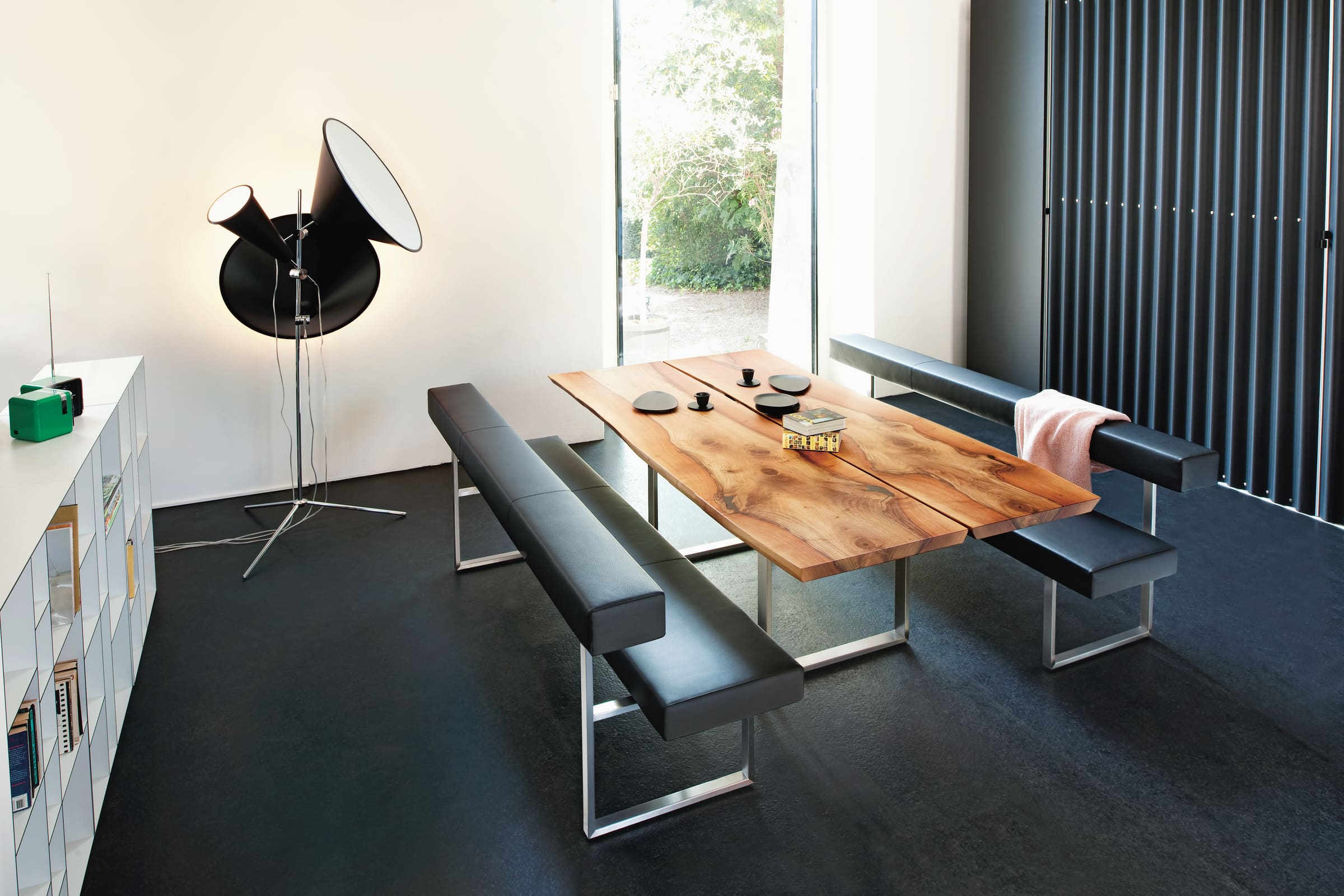 Moderner Essbereich mit Holztisch, schwarzen Sitzbänken der Girsberger Sitzbank Permesso, Stehlampe und einem Fenster, das Tageslicht einlässt. Moderner Essbereich mit Holztisch, schwarzen Sitzbänken der Girsberger Sitzbank Permesso, Stehlampe und einem Fenster, das Tageslicht einlässt.