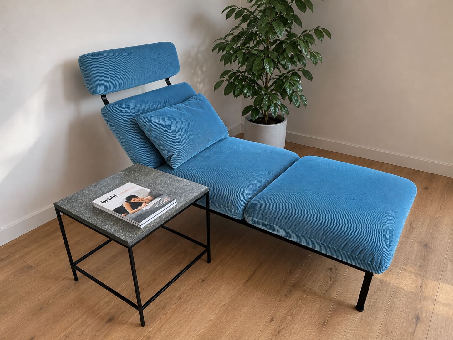 Die blaue Chaiselongue Liege Roro 98 steht neben einem kleinen Beistelltisch mit einer Zeitschrift, neben einer Topfpflanze auf einem Holzboden. Die blaue Chaiselongue Liege Roro 98 steht neben einem kleinen Beistelltisch mit einer Zeitschrift, neben einer Topfpflanze auf einem Holzboden.